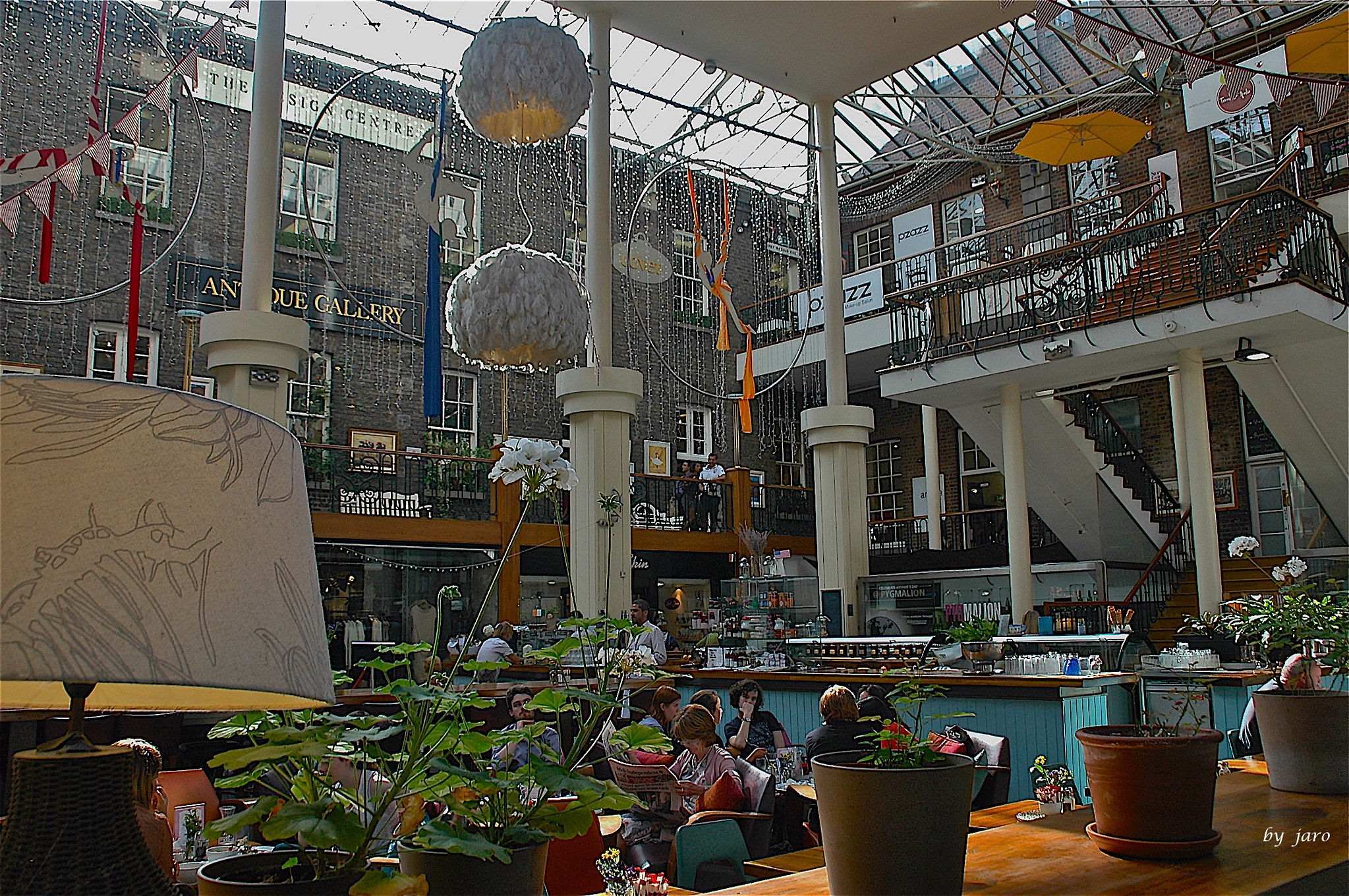 Photograph of people sitting in an outdoor cafe. There are potted plants in the foreground, and the entrance to an art gallery in the background.