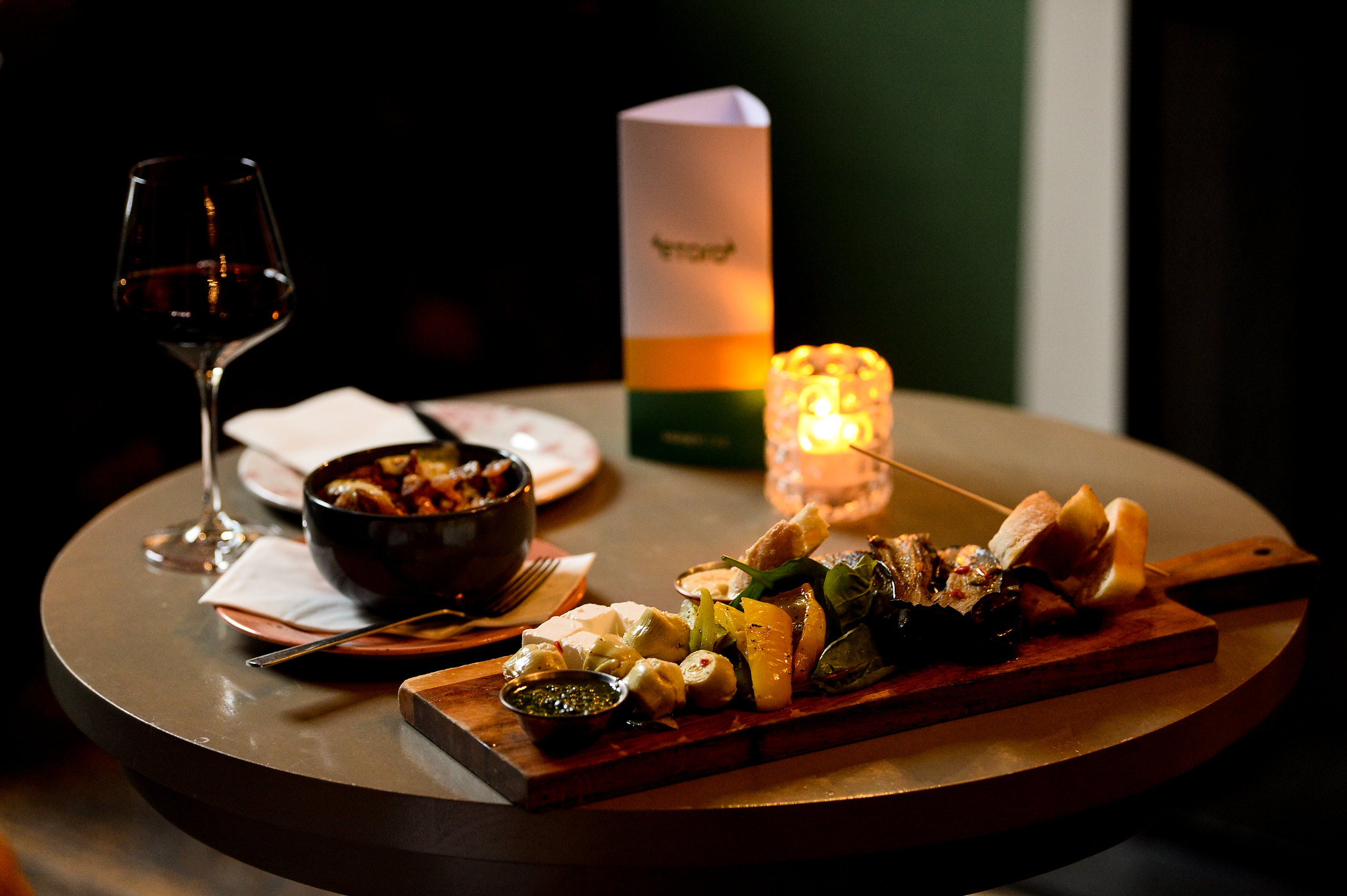Photograph of vegetables and cheese on a wooden board, alongside a small bowl and a glass of wine, all on a candlelit table.