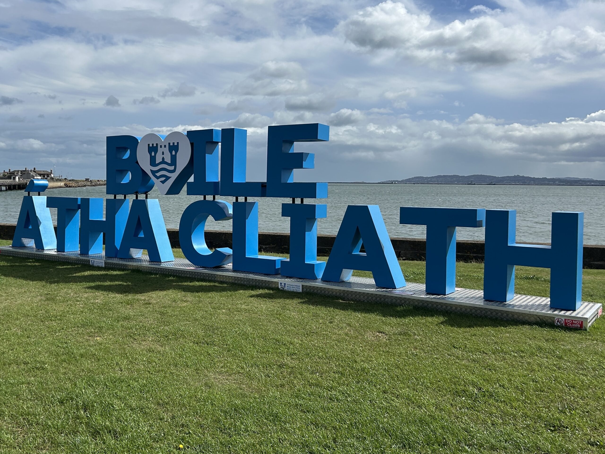 The Irish phrase 'Baile Átha Cliath' in large letters in a coastal park on a sunny day.