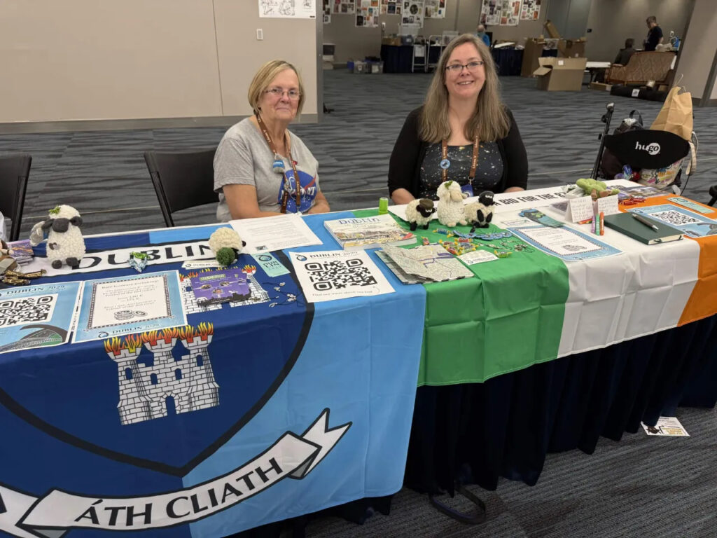 Two women at a table decorated with the Dublin and Irish flags and covered with Con paraphernalia. 