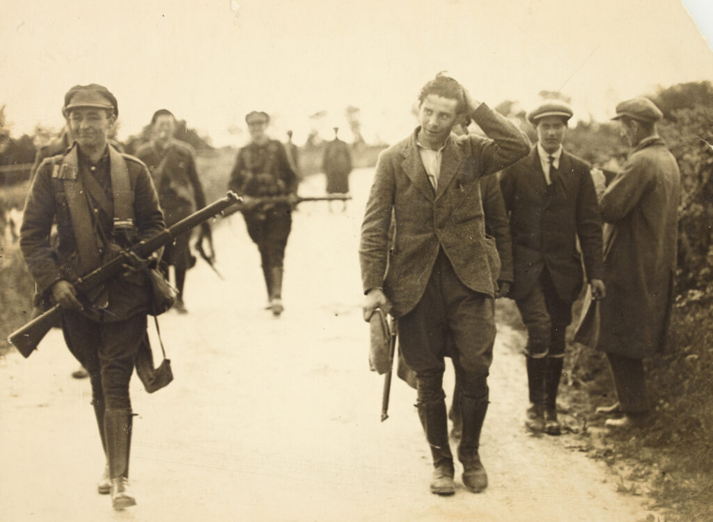 A black-and-white sepia photograph of armed soldiers escorting a man in civilian clothes.