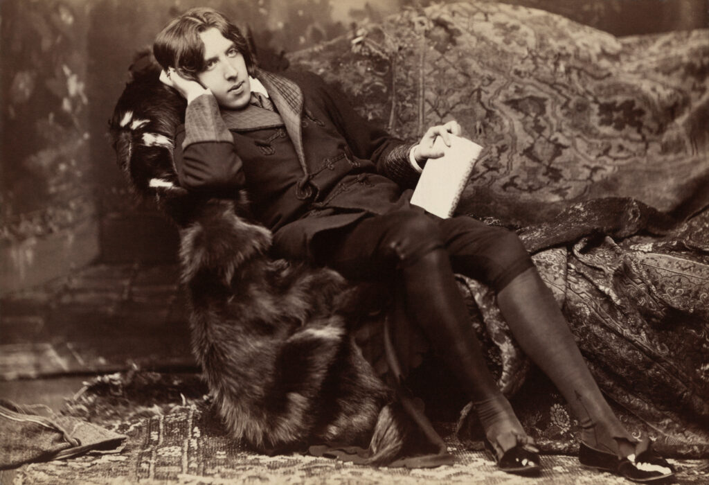 A black-and-white photo of very well-dressed man with a book of poetry in his hand reclining on a sofa covered in luxurious rugs. The man's style of dress is from the 19th Century.