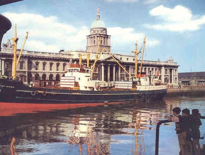 A photo from the 1950s showing an active dock outside the Custom House, a large Georgian building. A ship is positioned right in front of the building. The photo was taken from across the river and some children are looking at the boat from this side.