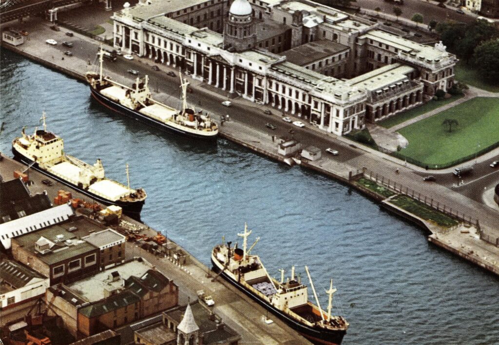 A busy docklands and industrial site from the 1950s. The Custom house is visible with a ship docked in front of it. Ships are also docked on the other side of the river.