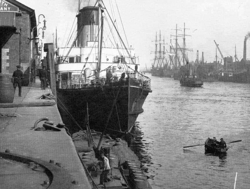 A Photograph of Dublin's docklands from the early 20th Centure. A steamship sits at the docks, with sail ships in the distance on the other side of the river.