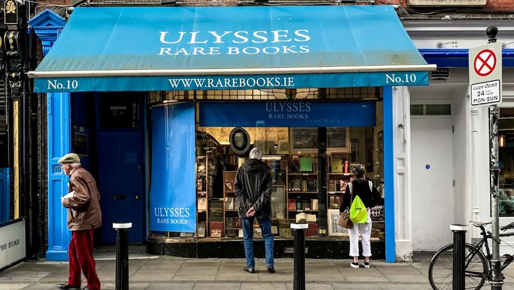 Photo of a bookshop with a bright blue awning and the name 'Ulysses Rare Books' in white. Two people are looking in the window and an elderly man is passing by.